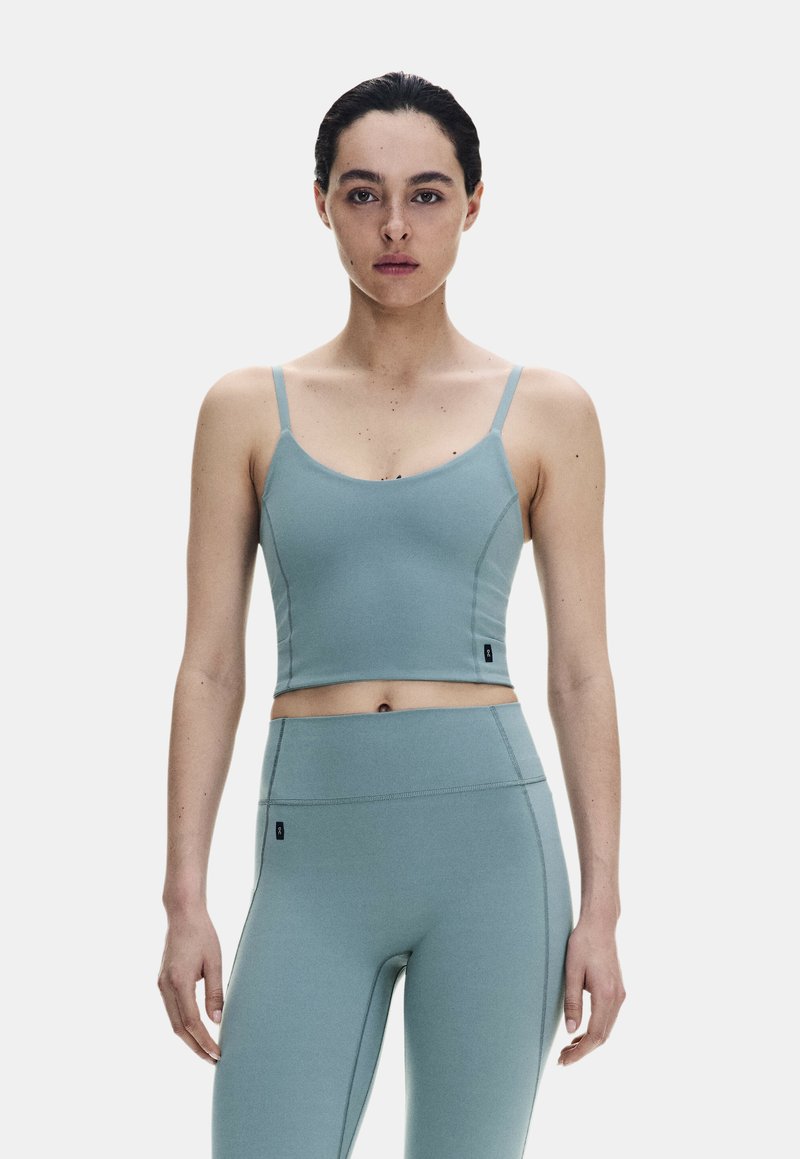 Jeune femme aux cheveux foncés attachés, porte un crop top bleu clair ajusté et un legging taille haute, debout devant un fond clair uni.