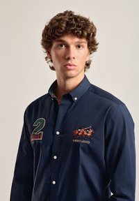 Camisa de botones color navy, de material de algodón, con el número "2" bordado y un logo de jinetes. Cuello interior de cuadros en contraste, corte regular.
