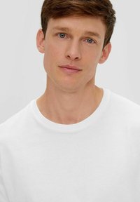 Weißes Baumwoll-T-Shirt mit rundem Halsausschnitt. Glatte Textur, taillierte Passform mit kurzen Ärmeln. Schlichte und minimalistische Ästhetik.