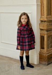 Vestido de cuadros con un cuello rojo, mangas largas y detalles con fruncido. Medias hasta la rodilla en color marino y zapatos oscuros completan el conjunto. Fondo de textura madera.