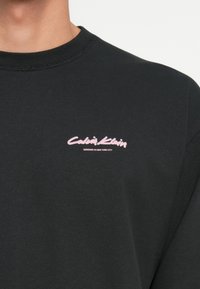 Schwarzer Crewneck-Sweatshirt aus Baumwolle, mit einem pinken gestickten "Calvin Klein"-Logo auf der linken Brust und "DESIGNED IN NEW YORK CITY."