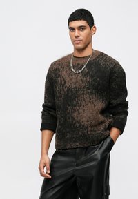 Barna és fekete mintás kötött pulóver kerek nyakkal és hosszú ujjakkal, fekete bőr nadrággal viselve. Lánc nyaklánc, mint kiegészítő.