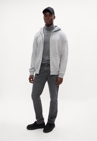 Felpa grigia con zip sopra un dolcevita grigio, abbinata a jeans grigio scuro e scarpe nere, caratterizzata da un design semplice e informale.