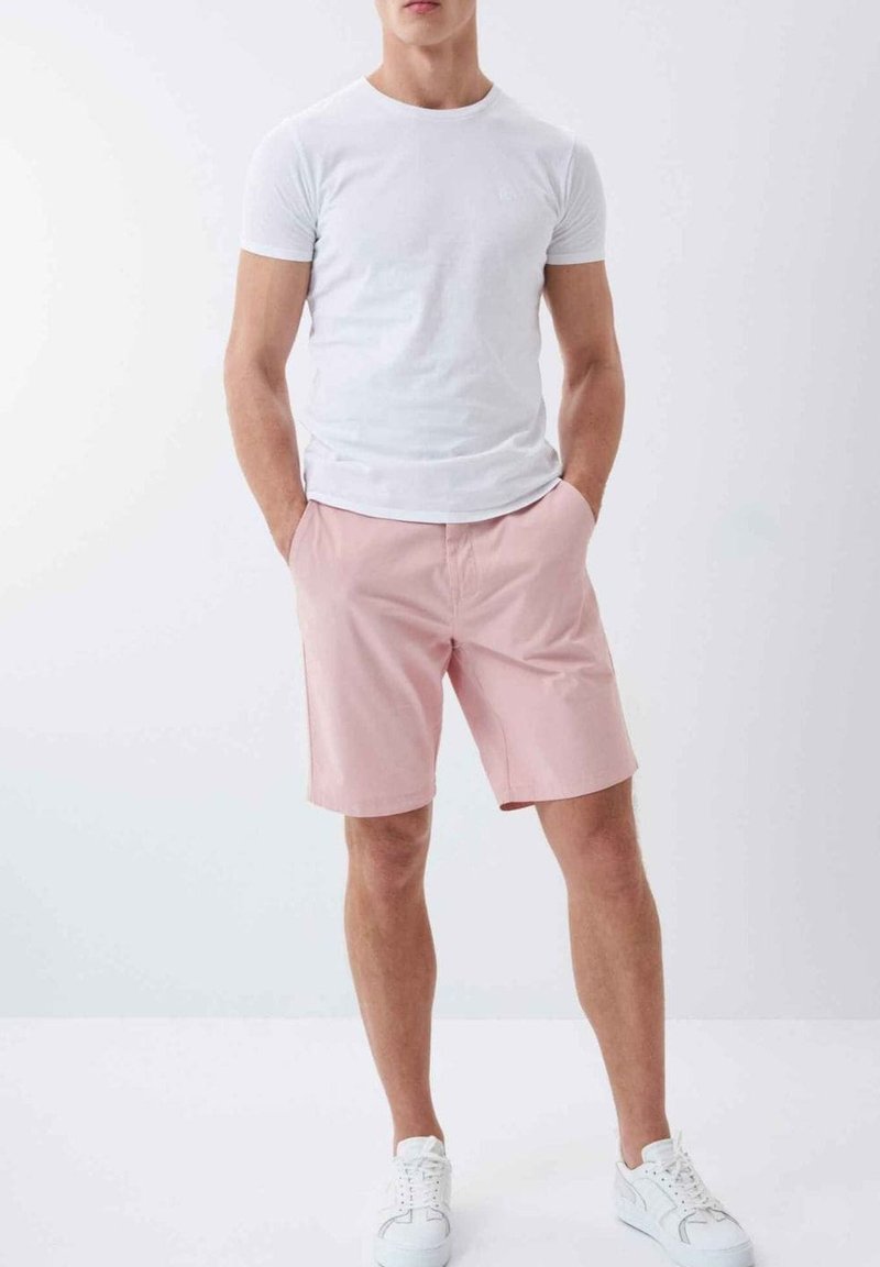 Camiseta blanca de manga corta combinada con pantalones cortos rosa pálido, con bolsillos frontales. Calzado incluye zapatillas blancas. Look sencillo y casual.