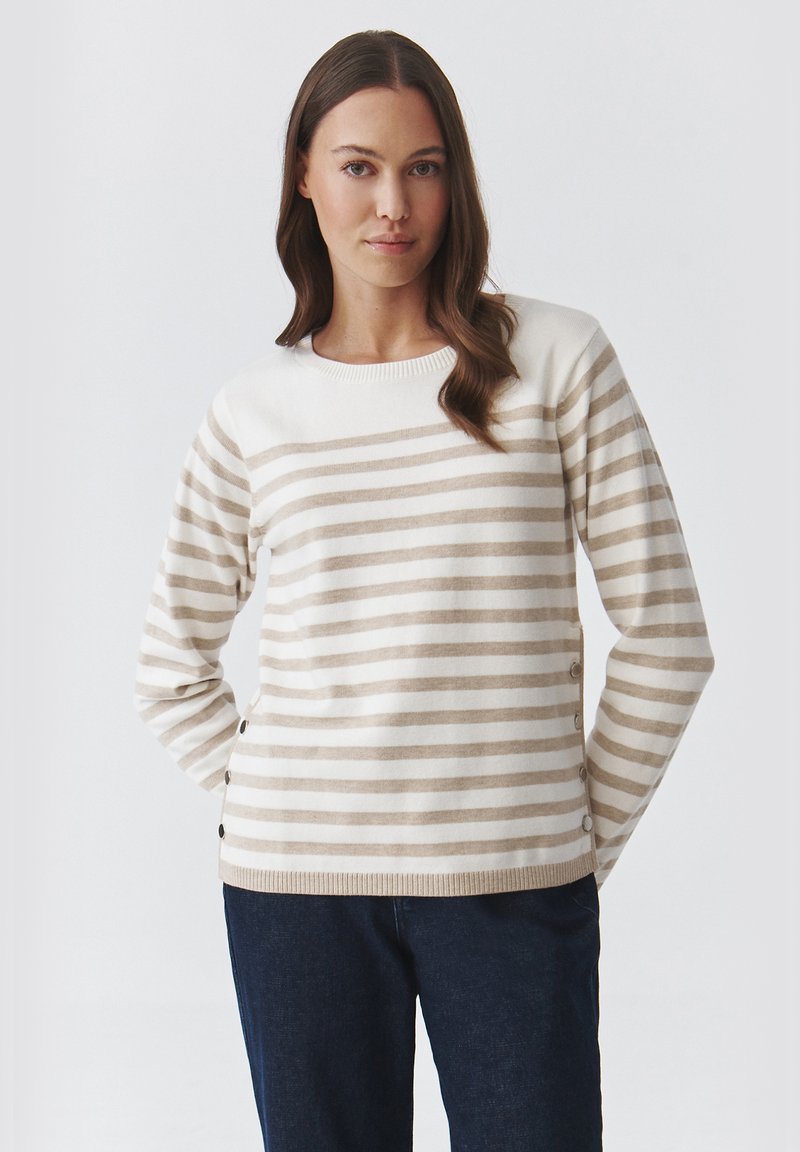 Cremfarbener Pullover mit beigen horizontalen Streifen, geripptem Halsausschnitt und Knopfdetails an der Seite. Getragen mit dunkelblauen Jeans.