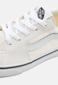 Bijele suede tenisice s bijelim vezicama, sivi kožni trak, platneni akcenti i crna gumena potplat. Vans logo na jeziku.