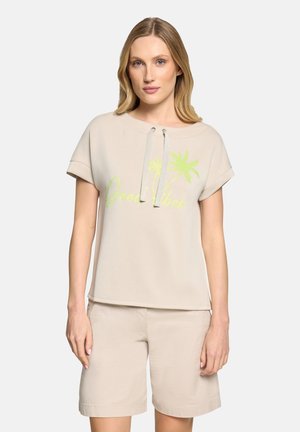 Lyshåret kvinde iført beige kortærmet hættetrøje med teksten "Good Vibes" og palmetræsgrafik, kombineret med beige shorts, stående mod en hvid baggrund.