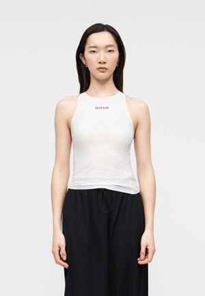 Donna giovane con capelli lisci e scuri che indossa un top bianco senza maniche Calvin Klein e pantaloni neri larghi, in piedi contro uno sfondo semplice.