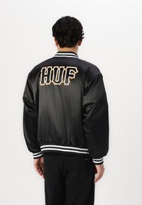 Jachetă din satin negru cu manșete și tiv în dungi albe, având un mare logo brodat "HUF" în portocaliu și alb pe spate.