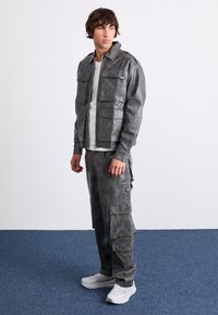 Veste grise en similicuir avec plusieurs poches à rabat, associée à un pantalon cargo assorti. Le mannequin porte un t-shirt blanc et des baskets blanches.