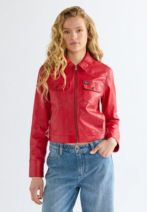 Jeune femme aux longs cheveux bouclés portant une veste en cuir rouge zippée et un jean bleu, debout avec une main dans la poche.