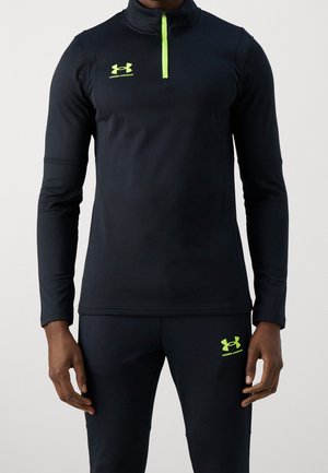 Man wearing black Under Armour long-sleeve top with neon yellow zipper and matching black pants with neon yellow logo.