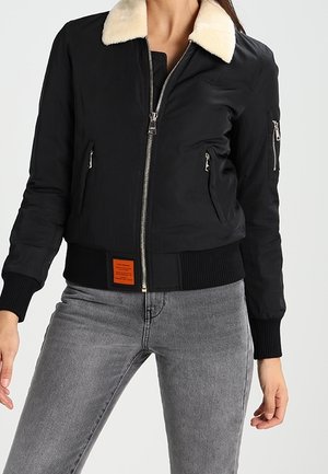 Kvinde iført sort lynlåsjakke med hvid pelskrave og grå jeans, stående mod en ensfarvet lys baggrund.