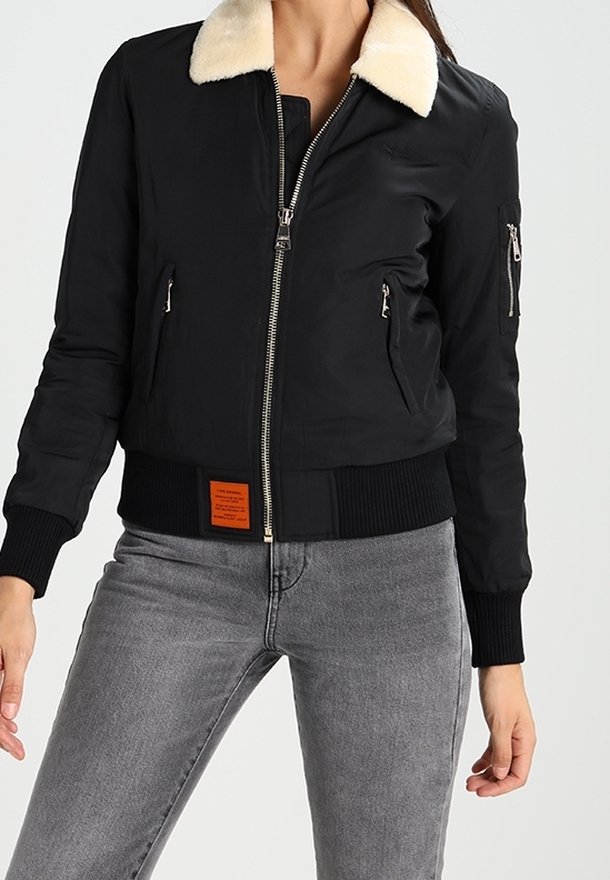 Femme portant une veste noire zippée avec un col en fourrure blanche et un jean gris, debout devant un fond clair uni.