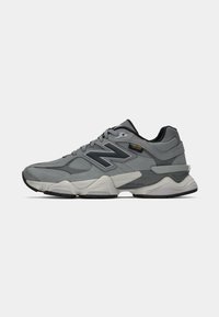 Graues Sport schuh mit einem Obermaterial aus verschiedenen Materialien, das Wildleder und Mesh umfasst, schwarzen Akzenten, dickem Sohlen und einem auffälligen 'N'-Logo an der Seite.