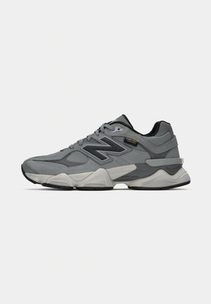 Scarpa sportiva grigia con tomaia in materiali misti, caratterizzata da suede e mesh, accenti neri, suola spessa e un logo 'N' prominente sul lato.