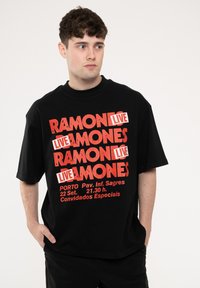 Black short-sleeve t-shirt featuring bold red text that reads "RAMONES LIVE" and event details. Material appears soft with a relaxed fit.