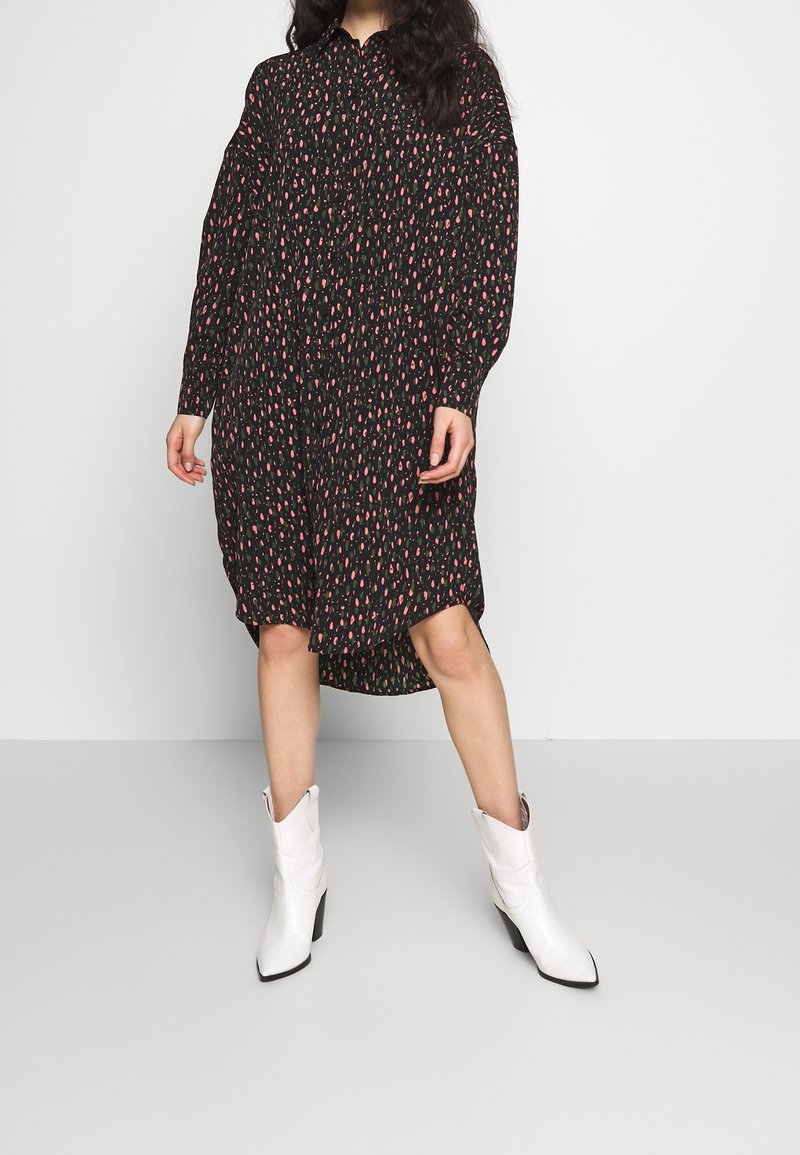 Femme portant une robe midi noire à manches longues avec un motif rose et des bottines blanches, debout devant un fond gris clair uni.
