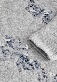 Grau strukturiertes Gewebe mit verstreuten Pailletten in Silber- und Blautönen. Verfügt über einen umgeschlagenen Ärmelaufschlag für zusätzlichen Detail.