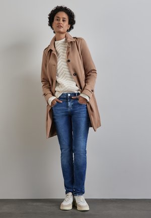 Gabardina beige sobre un suéter crema con un patrón de zigzag, jeans azules y zapatillas blancas. Fondo liso, manos en los bolsillos.