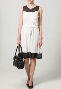 Robe blanche avec un haut transparent noir et un ourlet noir, cintrée à la taille. Associée à un sac à main noir et des chaussures noires à bout fermé.