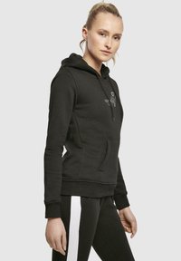 Haut à capuche noir avec une poche frontale, cordon de serrage et détail de logo. Fabriqué en tissu doux, avec une coupe ajustée et des poignets côtelés.