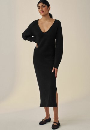 Abito nero a coste con scollatura profonda a V e maniche lunghe, dotato di spacchi laterali e silhouette aderente. Abbinato a ballerine nere.