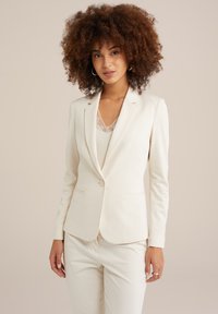 Blazer en ivoire avec une fermeture �à un bouton, coupe ajustée et revers crantés. Texture lisse, associé à un pantalon assorti.