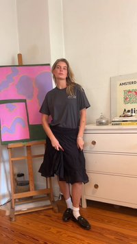 Tricou gri cu un logo, fustă neagră cu un design fluid, sosete albe și mocasini negri; pictură abstractă colorată în fundal.