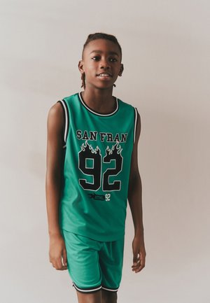 Junge trägt eine grüne ärmellose Sportbekleidung mit "SAN FRAN 92" vorne aufgedruckt und steht vor einem schlichten weißen Hintergrund.