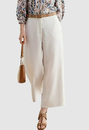 Mujer con pantalones anchos blancos, cinturón marrón con tachuelas y sandalias marrones con tachuelas a juego, sosteniendo un bolso marrón claro.