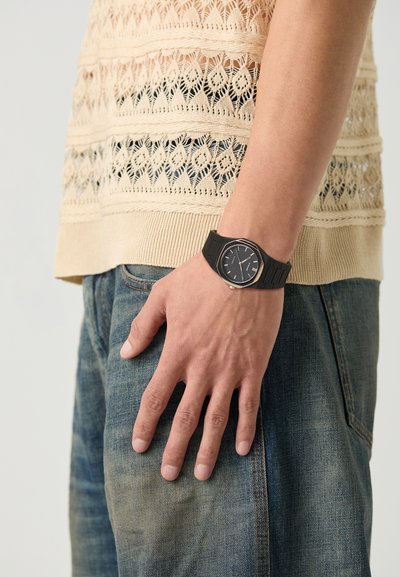 Montre noire avec un cadran rond, bracelet en silicone noir, portée sur une main. Tenue décontractée comprenant un haut en tricot beige et un jean bleu délavé.