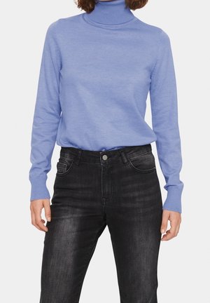 Persona que lleva un suéter de cuello alto azul claro y vaqueros negros de cintura alta, de pie con los brazos relajados a los lados sobre un fondo blanco.