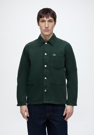 Junger Mann trägt eine dunkelgrüne, mit Knöpfen versehene Jacke mit Kragen und drei vorderen Taschen, steht vor weißem Hintergrund.