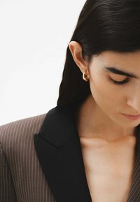 Blazer marrón a rayas con cuello negro, combinado con pendientes de aro dorados. La modelo tiene el cabello oscuro y está mirando hacia abajo.