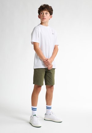 T-shirt en coton blanc, shorts vert olive, baskets blanches avec des accents verts, et chaussettes blanches rayées de bleu, debout les bras croisés.