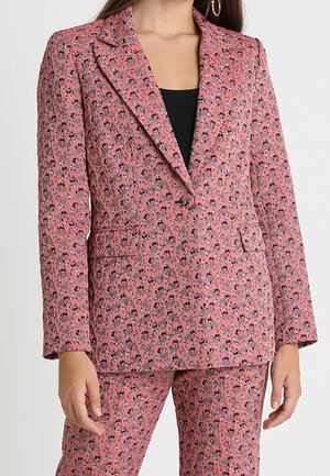 Kobieta ubrana w dopasowaną różową marynarkę w kwiaty i pasujące spodnie, do tego czarna bluzka pod spodem.