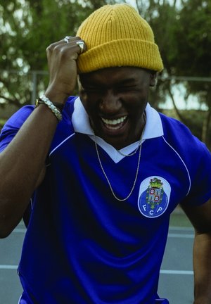 Blauwe sporttrui met witte accenten en FCP-logo, gele gebreide muts, zilveren ketting en armbanden. Onderwerp glimlachend en gebarend.