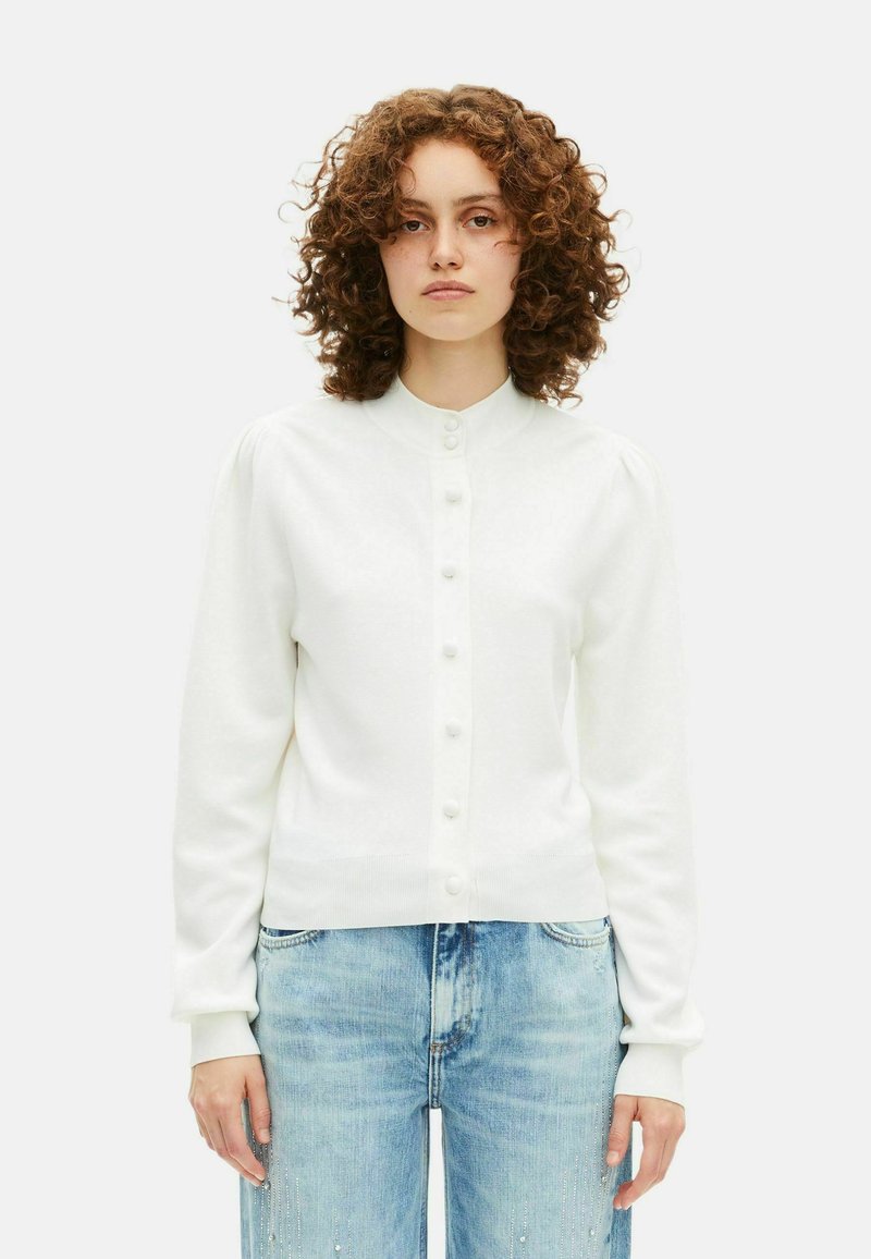 Femme aux cheveux bouclés portant un cardigan blanc à boutons et un jean bleu clair avec de subtils ornements sur les cuisses.