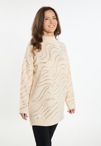 Beiger Pullover mit hohem Kragen, der metallische Wellenmuster und eine weiche Textur aufweist. Kombiniert mit schwarzen, eng anliegenden Hosen.