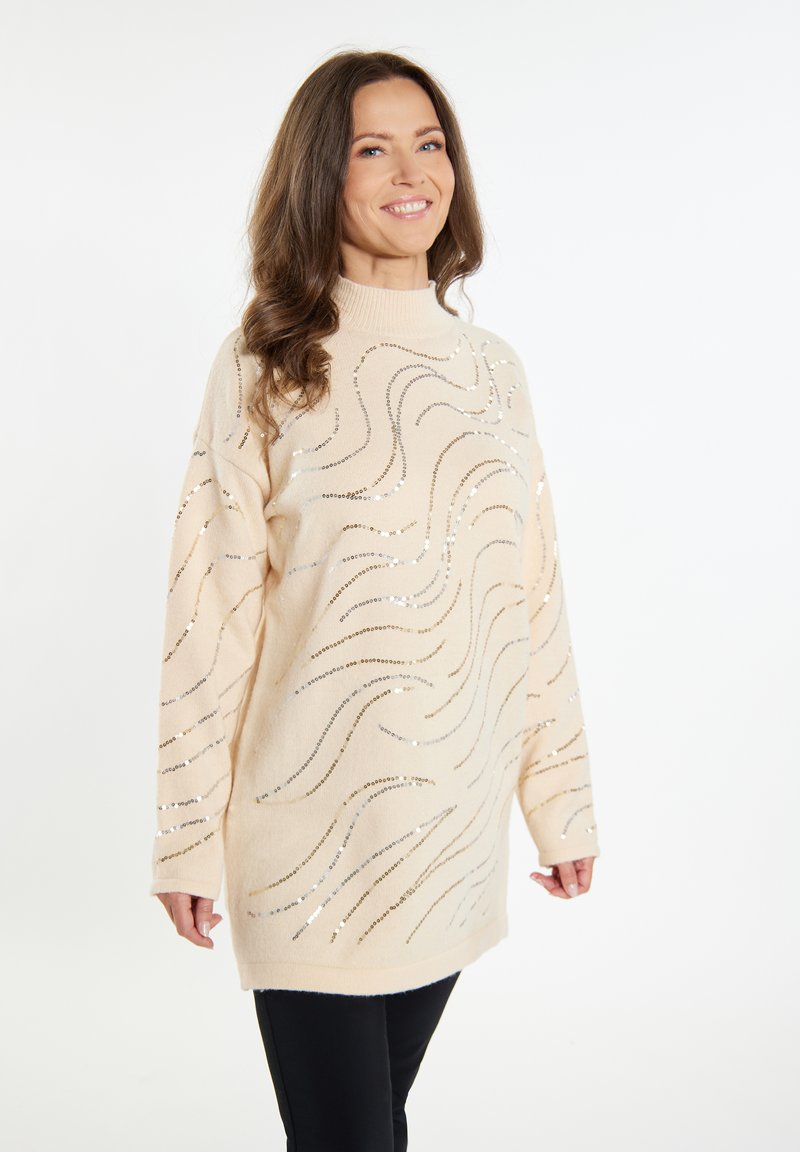 Beiger Pullover mit hohem Kragen, der metallische Wellenmuster und eine weiche Textur aufweist. Kombiniert mit schwarzen, eng anliegenden Hosen.