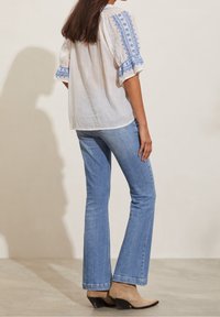 Vit texturerad blus med blå brodyr på ärmarna, kombinerad med ljusblå utsvängda jeans och bruna ankelstövlar. Enkla, avslappnade design.