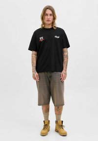 Hombre con tatuajes que lleva una camiseta gráfica negra, pantalones cortos grises largos, calcetines beige y botas de trabajo color canela, de pie contra un fondo claro y liso.