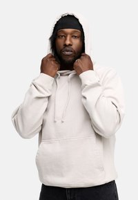 Hellbeige Kapuzenpullover aus Baumwollmischgewebe, mit einer Fronttasche, Kordelzugkapuze und Rippbündchen. Getragen von einer Person, die die Kapuze hält.