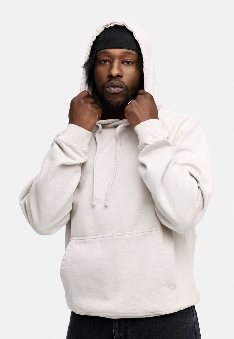 Hellbeige Kapuzenpullover aus Baumwollmischgewebe, mit einer Fronttasche, Kordelzugkapuze und Rippbündchen. Getragen von einer Person, die die Kapuze hält.