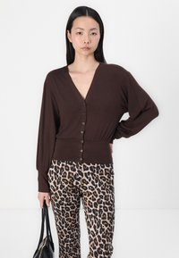 Brun langermet cardigan med V-hals og seks knappelukkinger, kombinert med leopardmønstrede bukser og en svart håndveske.