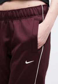 Burgundske sportske hlače s pojasom, bijelim bočnim trakama i vidljivim džepom. Na lijevoj strani nalazi se bijeli Nike logotip.