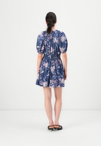 Femme aux cheveux bruns en chignon bas, portant une robe bleu à fleurs avec des manches bouffantes et des chaussures noires à talons bas, se tenant contre un fond blanc.