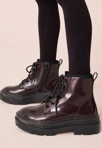 Bottes de cheville en cuir verni bordeaux avec bouts ronds, semelles en caoutchouc noir, devant à lacets et détail de fermeture éclair sur le côté. Col élastique noir.