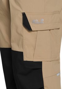 Μπεζ και μαύρο παντελόνι εξωτερικού χώρου με πλαϊνή τσέπη με καπάκι που φέρει το λογότυπο Jack Wolfskin σε γκρι χρώμα.